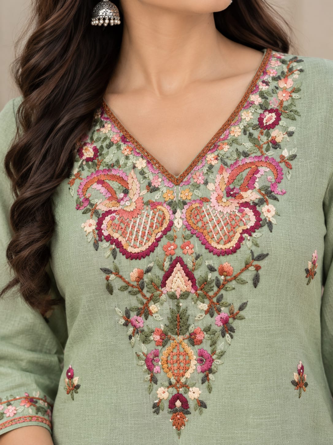 A close-up of a woman wearing a sage green embroidered kurta. The garment features intricate floral and paisley embroidery in pink, orange, red, and green around the V-neckline and on the front panel.