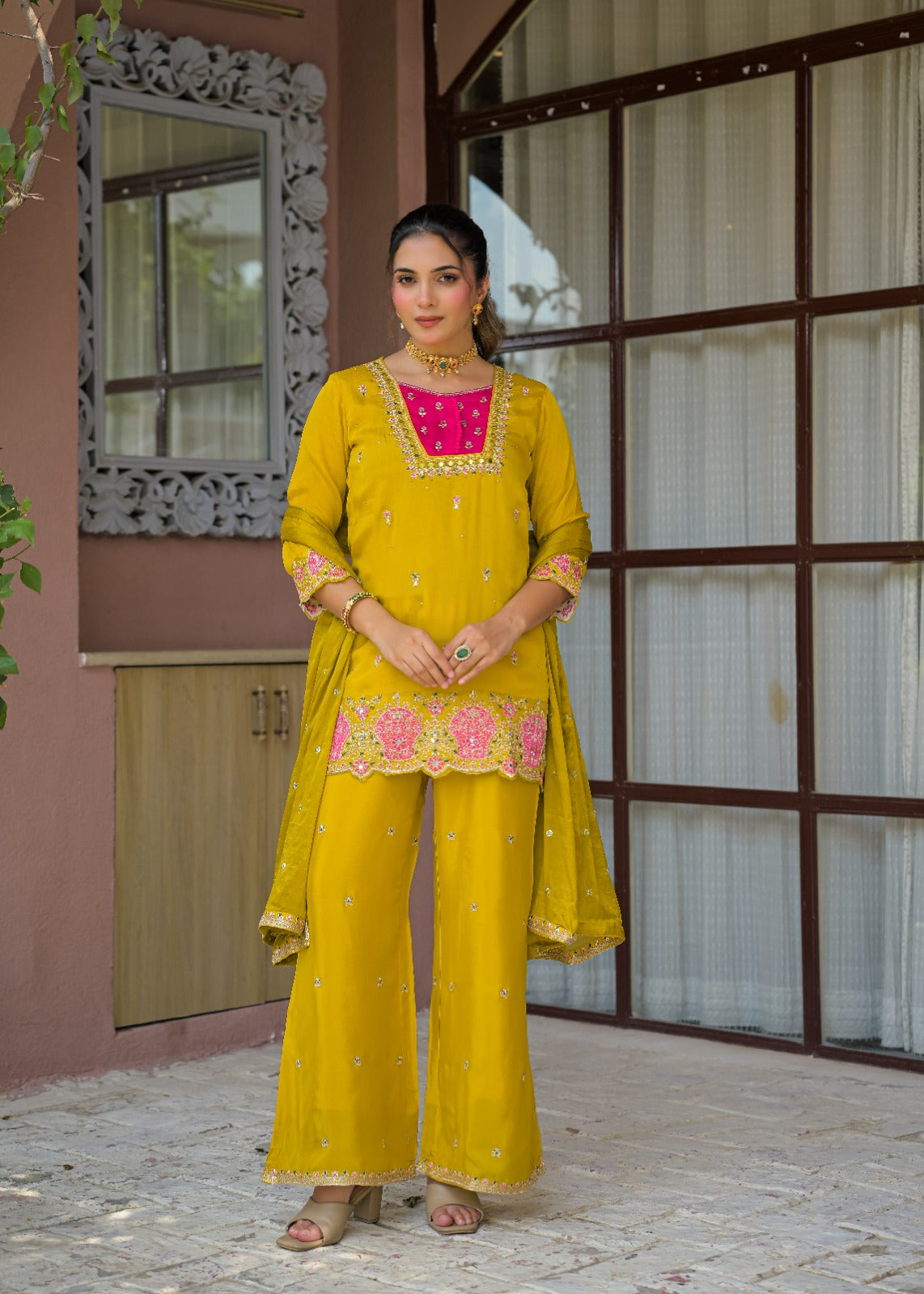 A woman stands outdoors wearing a bright yellow traditional outfit with pink floral embroidery and gold detailing, paired with matching wide-leg pants and a sheer dupatta.