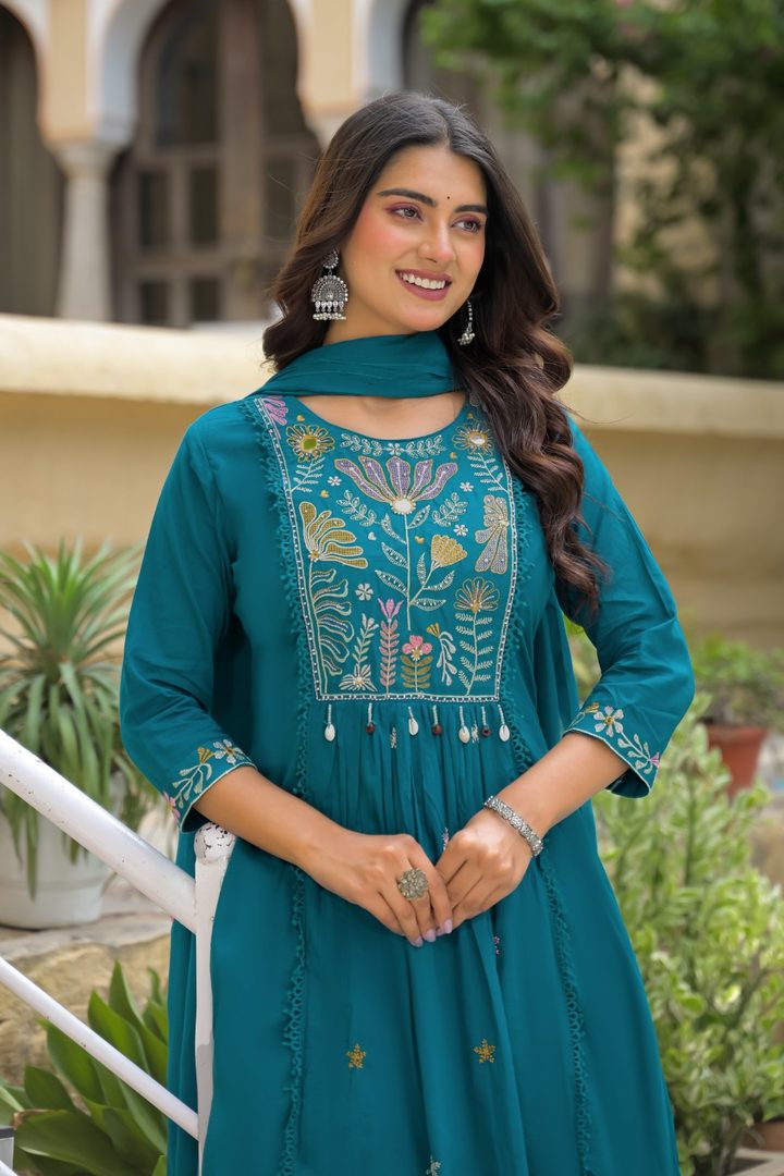 A woman wearing a teal green embroidered kurta with a matching dupatta, standing in an outdoor courtyard setting.