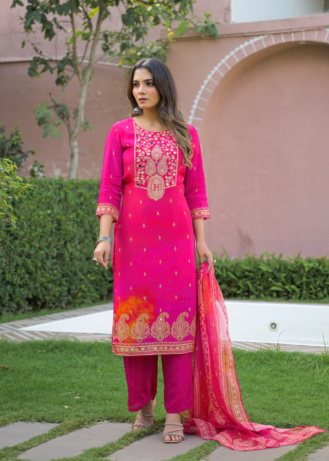 A woman stands outdoors on a garden path wearing a bright pink traditional outfit with gold embroidery, matching pants, and a sheer pink dupatta, with a pink stucco wall and green hedges in the background.