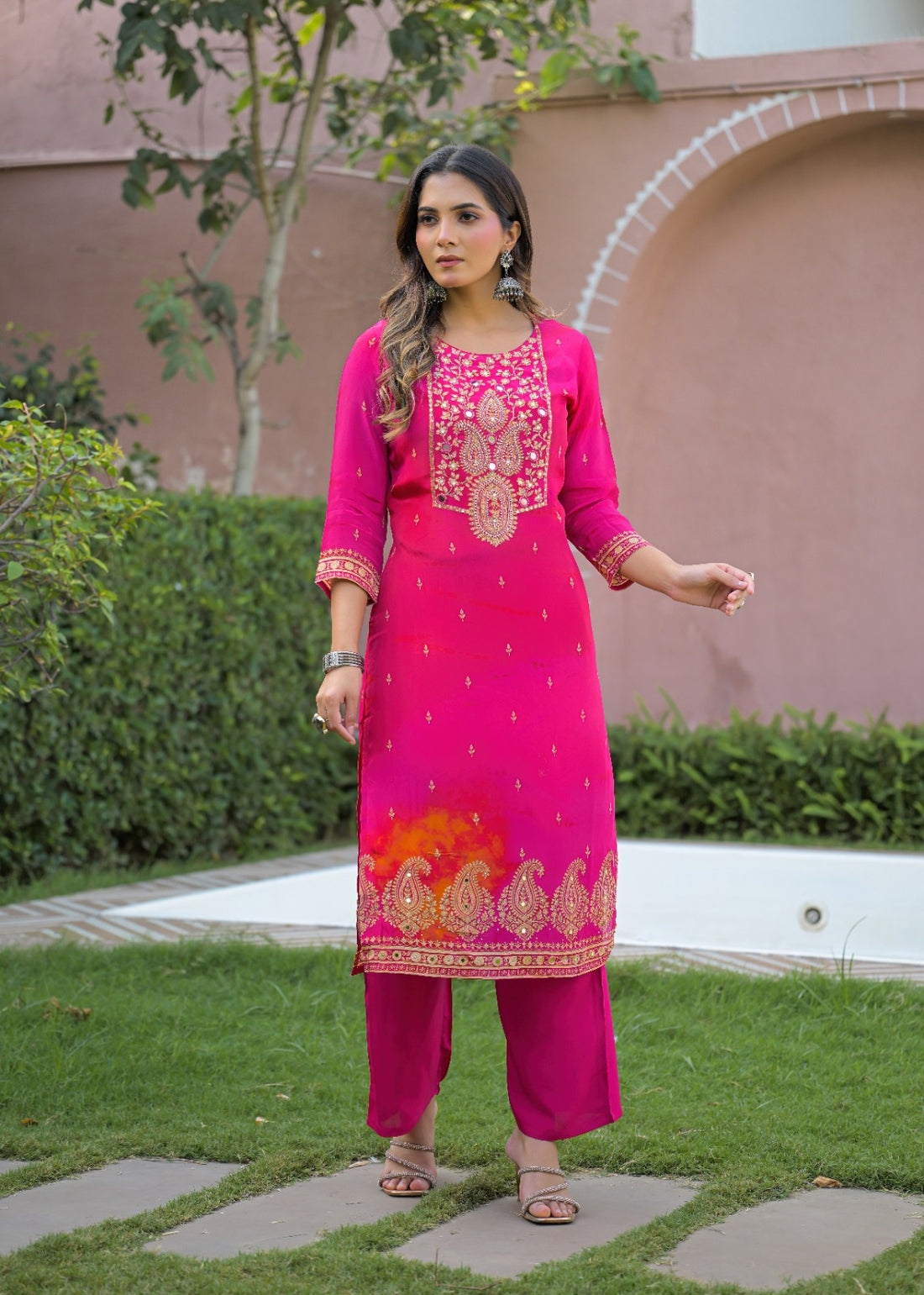 A woman stands outdoors on a garden path wearing a bright pink traditional outfit with gold embroidery, matching pants, and a sheer pink dupatta, with a pink stucco wall and green hedges in the background.