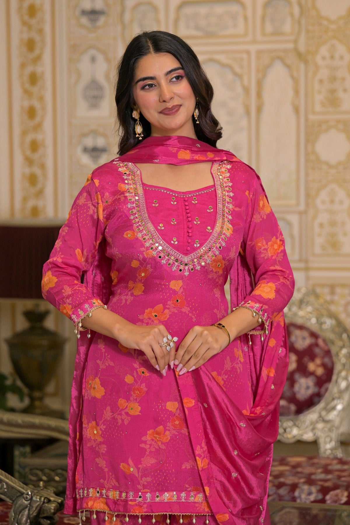 A woman modeling a bright pink traditional Indian salwar kameez suit with floral patterns, a matching dupatta, and ornate silver embroidery on the neckline, set against an ornate indoor background.