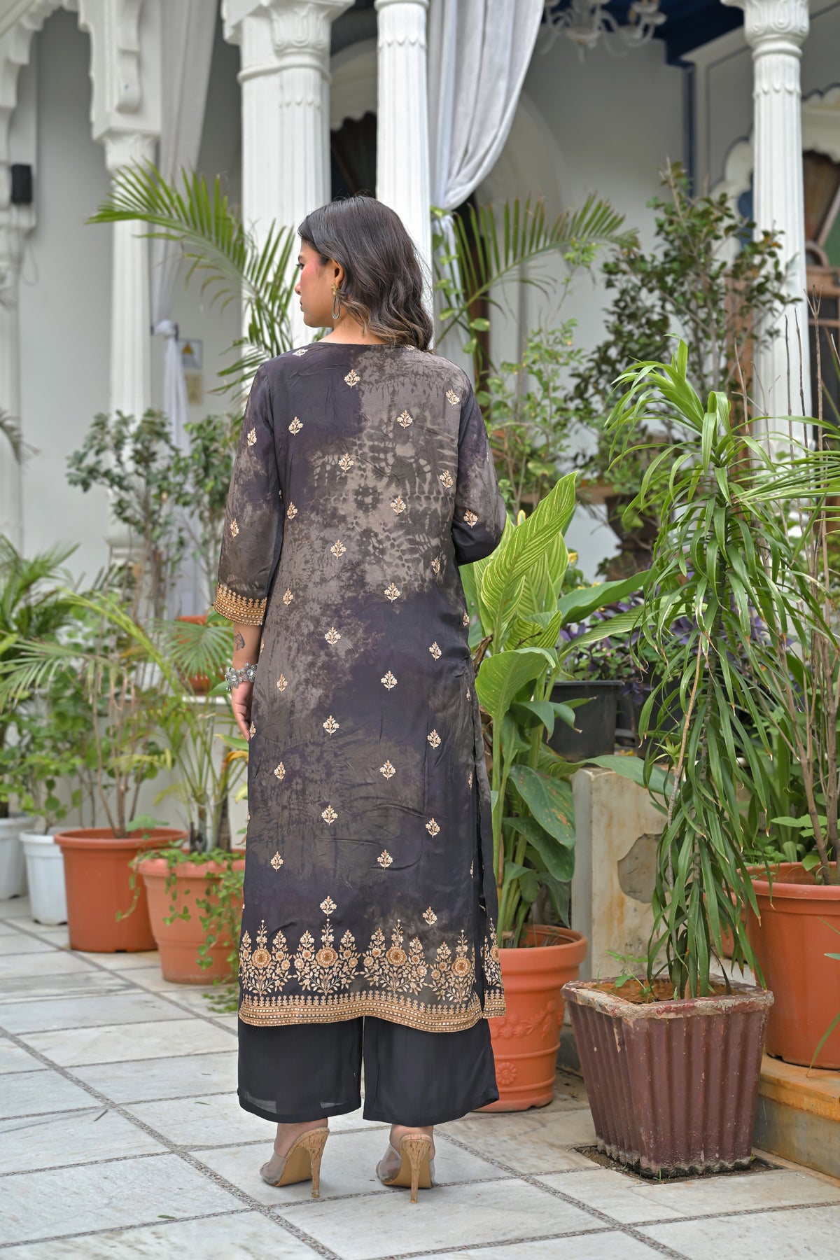 A woman stands with her back to the camera wearing a dark charcoal gray three-piece salwar kameez suit with gold embroidery, set against a backdrop of white pillars and potted plants.