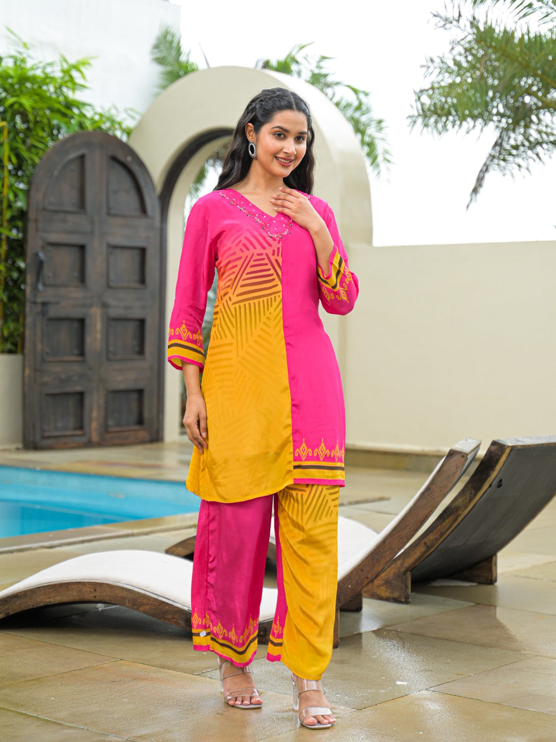 A woman modeling a bright pink and yellow traditional Indian outfit with matching pants, standing outdoors near a pool and lounge chairs.