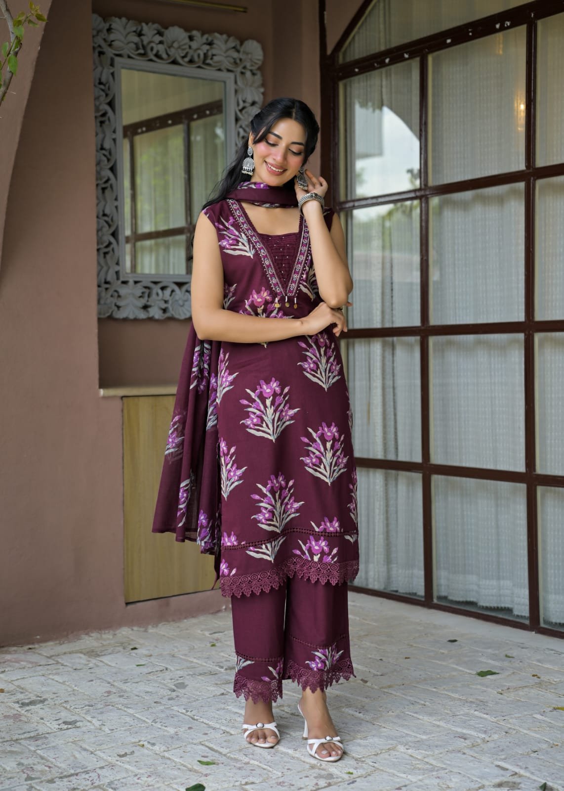 A woman models a three-piece traditional Indian salwar kameez suit in deep maroon with light pink floral embroidery, standing in front of a building with brown-framed glass windows.