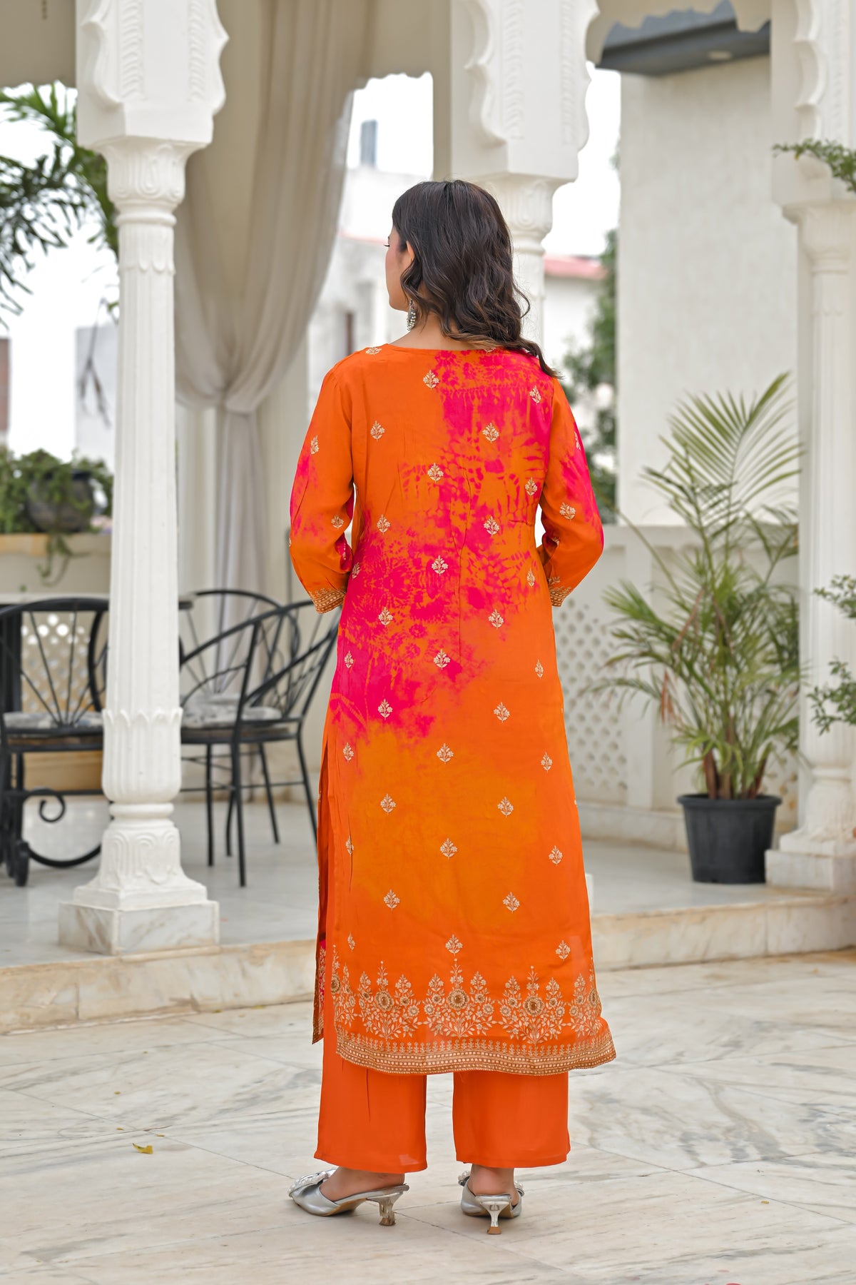 A woman viewed from behind wearing a bright orange traditional outfit with a large pink floral print on the back, paired with matching orange pants, standing in a courtyard with white pillars.