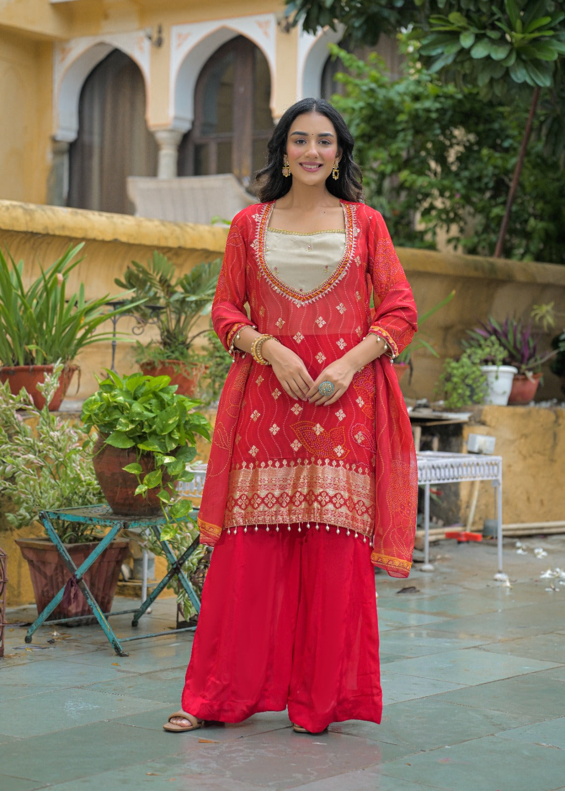 A woman stands outdoors wearing a bright red Indian ethnic outfit with a beige inner top, red embroidered kurta, matching wide-leg pants, and a red dupatta. The setting features a courtyard with potted plants, a stone floor, and a beige building in the background.