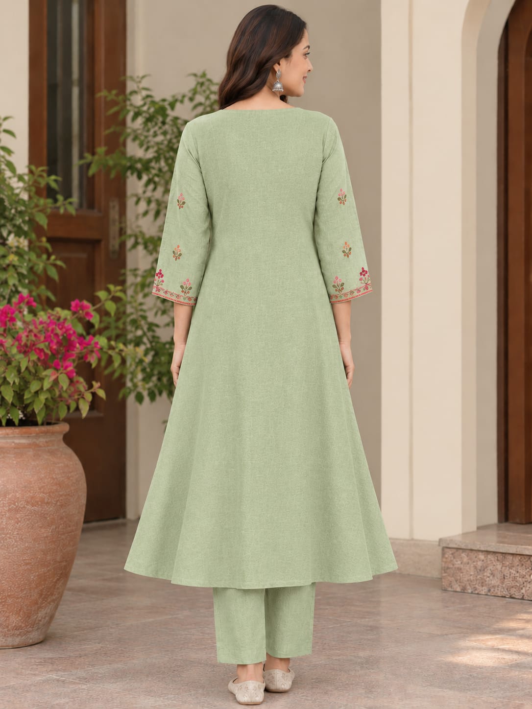 A woman is shown from the back wearing a light sage green kurta with three-quarter sleeves and floral embroidery on the cuffs, paired with matching pants. She is standing in front of a beige building with a large terracotta pot of pink flowers on the left.