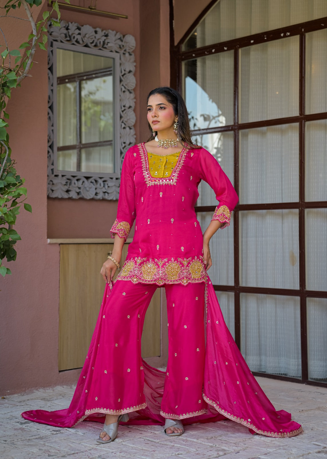A woman models a bright pink traditional three-piece salwar kameez suit with gold embroidery, standing in front of a pink stucco building with a decorative window frame.