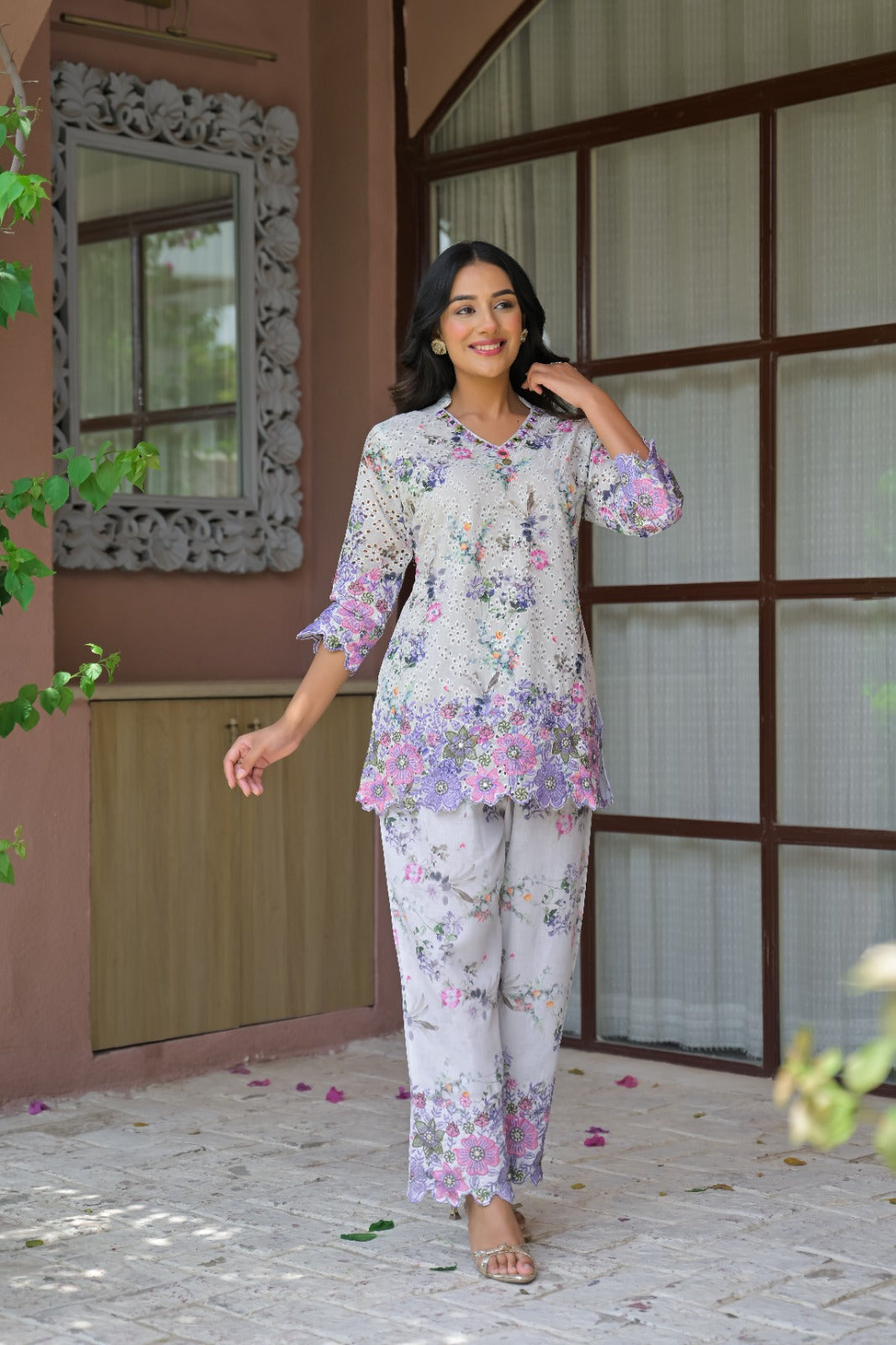 A woman modeling a two-piece floral pajama set with a light gray base and pink, purple, and green floral print, standing on a stone patio in front of a brown wooden window.