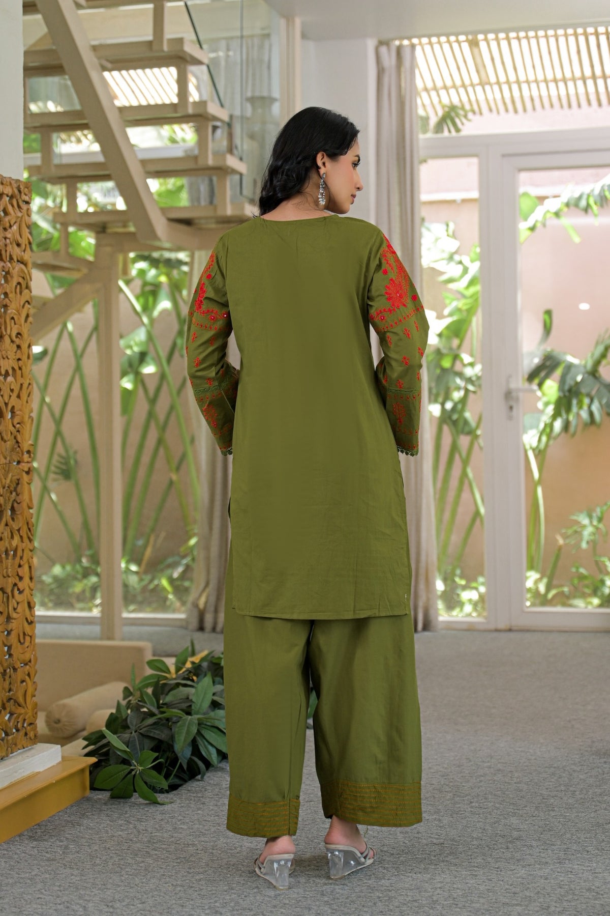 A woman wearing an olive green three-piece salwar kameez suit with red floral embroidery on the sleeves, shown from the back in a modern indoor setting with a staircase and glass doors.