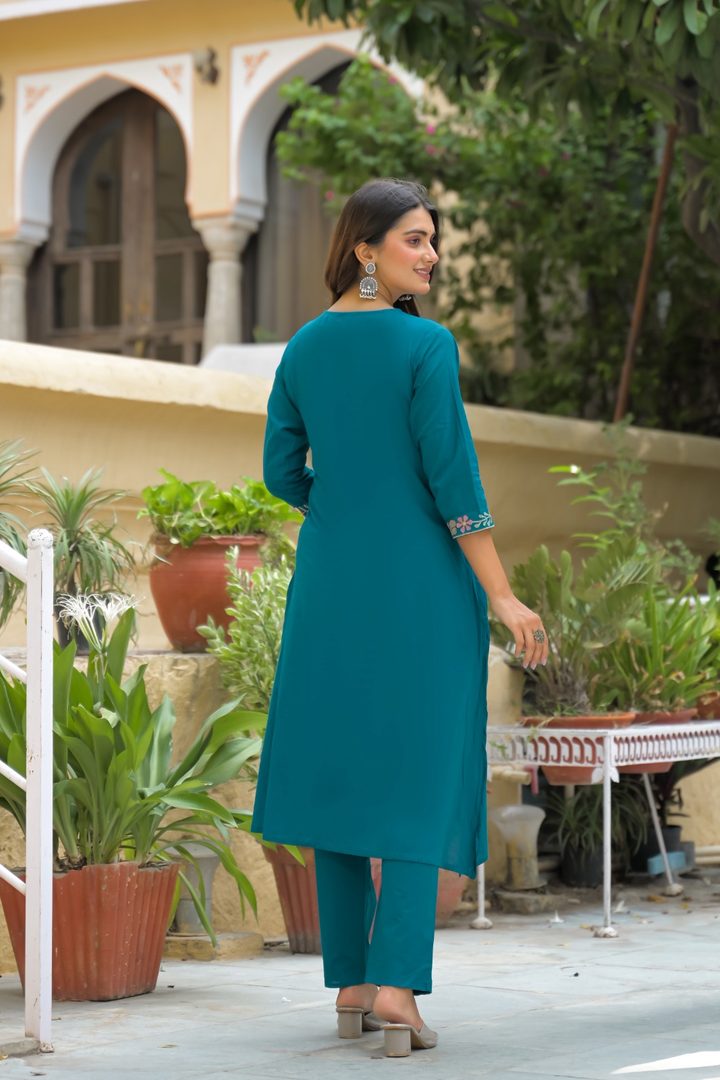 A woman wearing a teal green kurta and matching trousers stands outdoors in front of a building with arches and potted plants, photographed from the back.