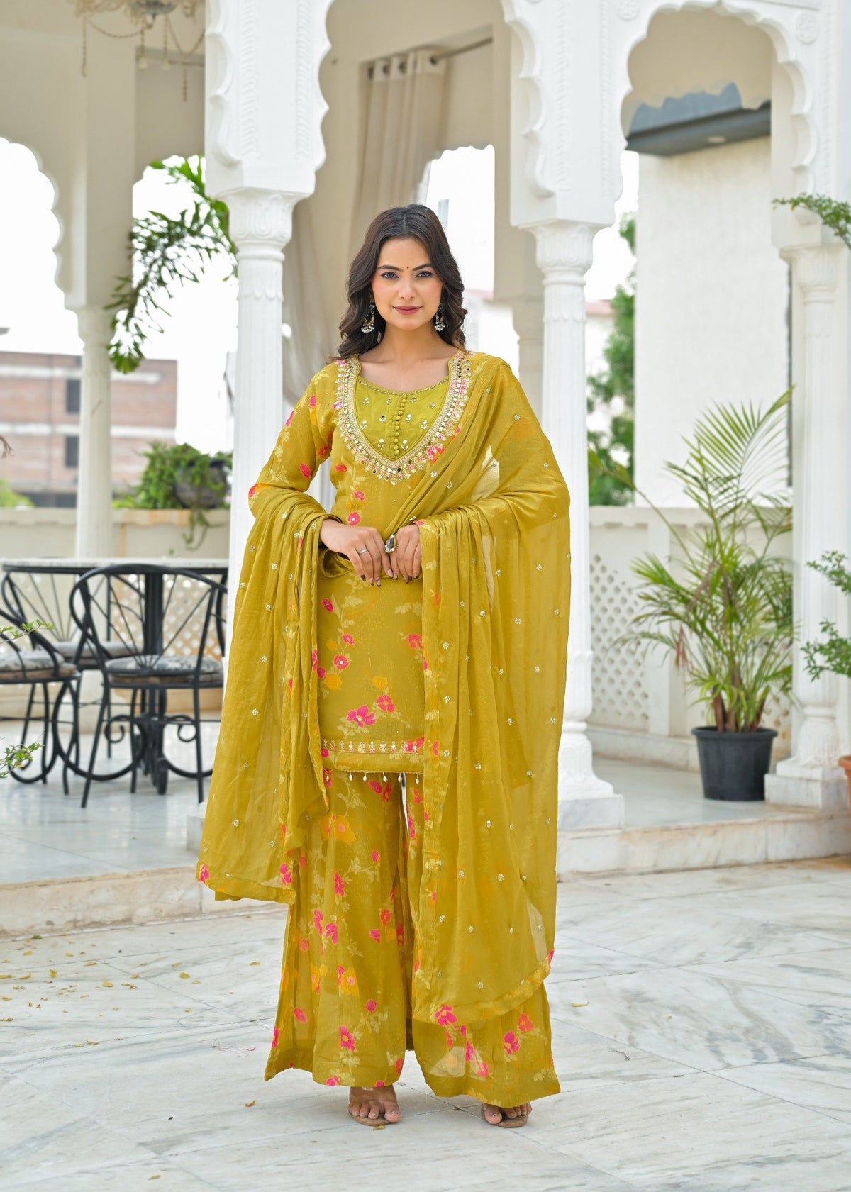 A woman models a yellow-green ethnic salwar kameez suit with pink floral embroidery and gold zari details, standing in a courtyard with white arches.