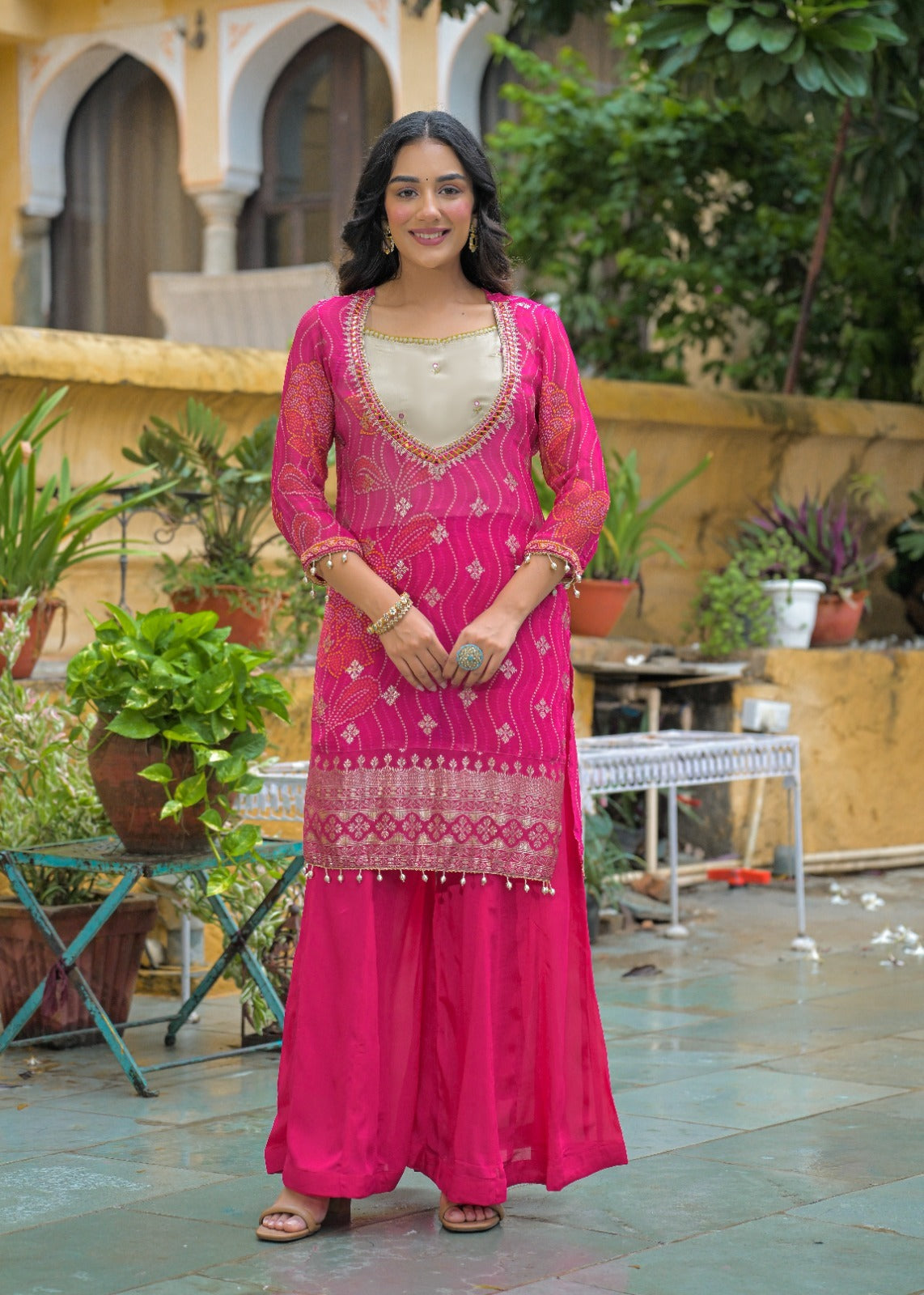 A woman stands outdoors wearing a bright pink traditional outfit with a cream inner top and detailed gold embroidery. The setting features a courtyard with potted plants, stone flooring, and a building in the background.