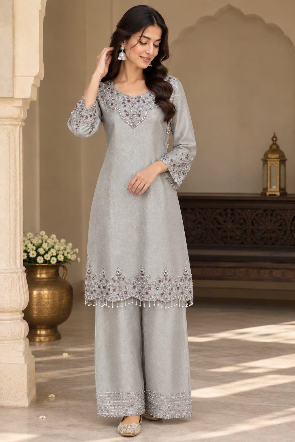 A woman modeling a light gray three-piece Indian ethnic suit with silver embroidery, standing in a traditional courtyard setting.