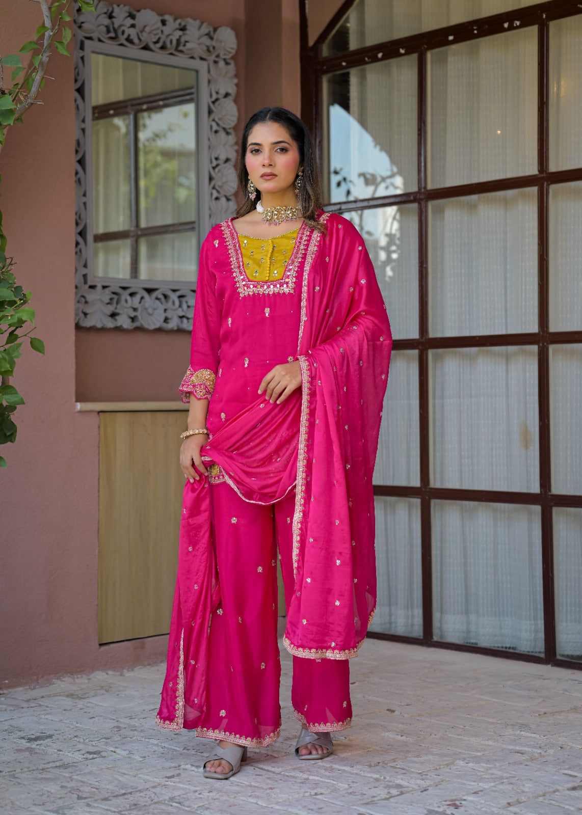 A woman models a bright pink traditional Indian outfit with a matching dupatta, featuring gold embroidery and a yellow embroidered neckline, standing in front of a pink stucco building with a large window.