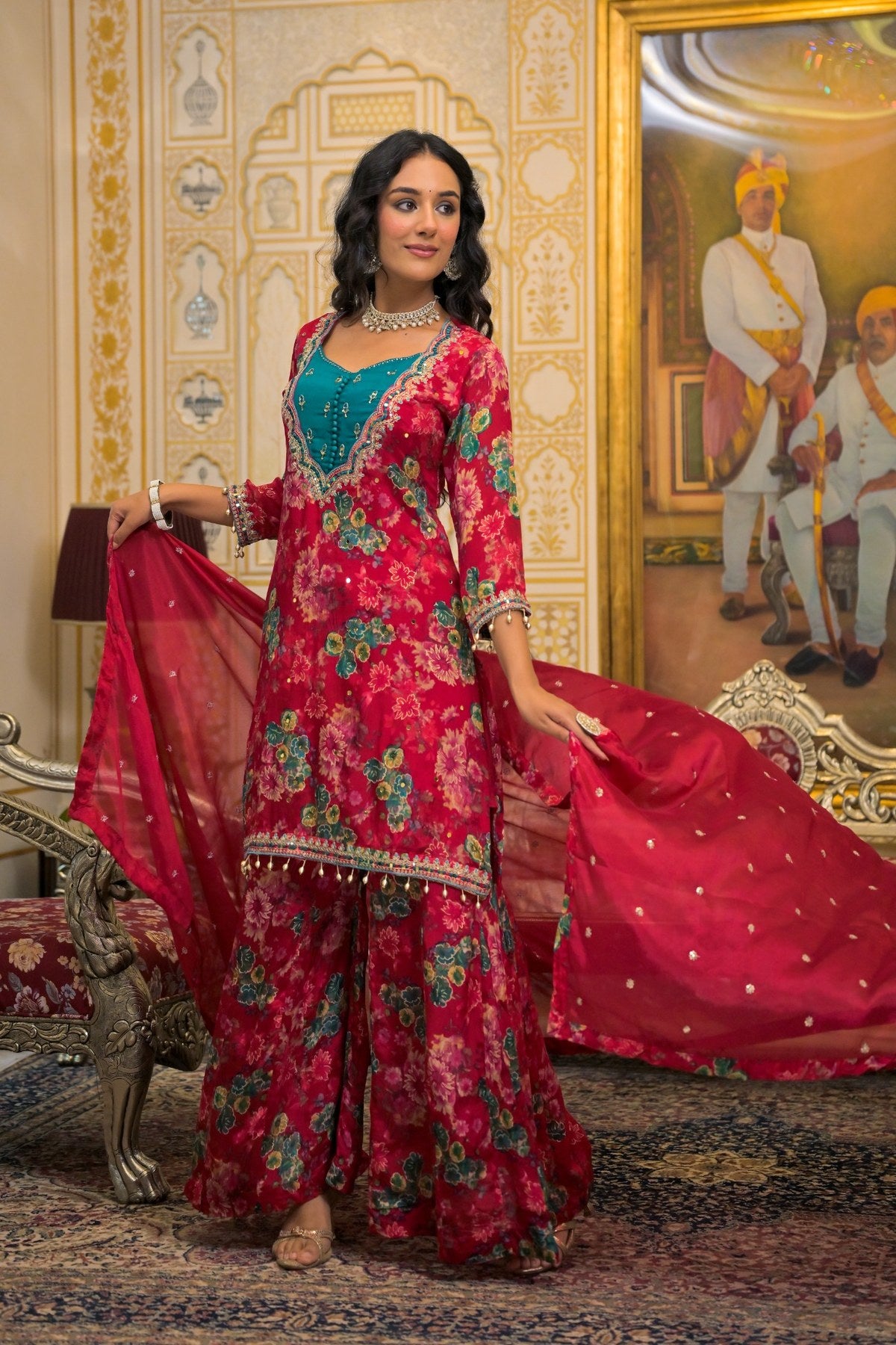 A woman models a traditional Indian salwar kameez outfit in a rich magenta floral print with a teal blue inner lining and a matching red dupatta, set against an ornate palace interior.