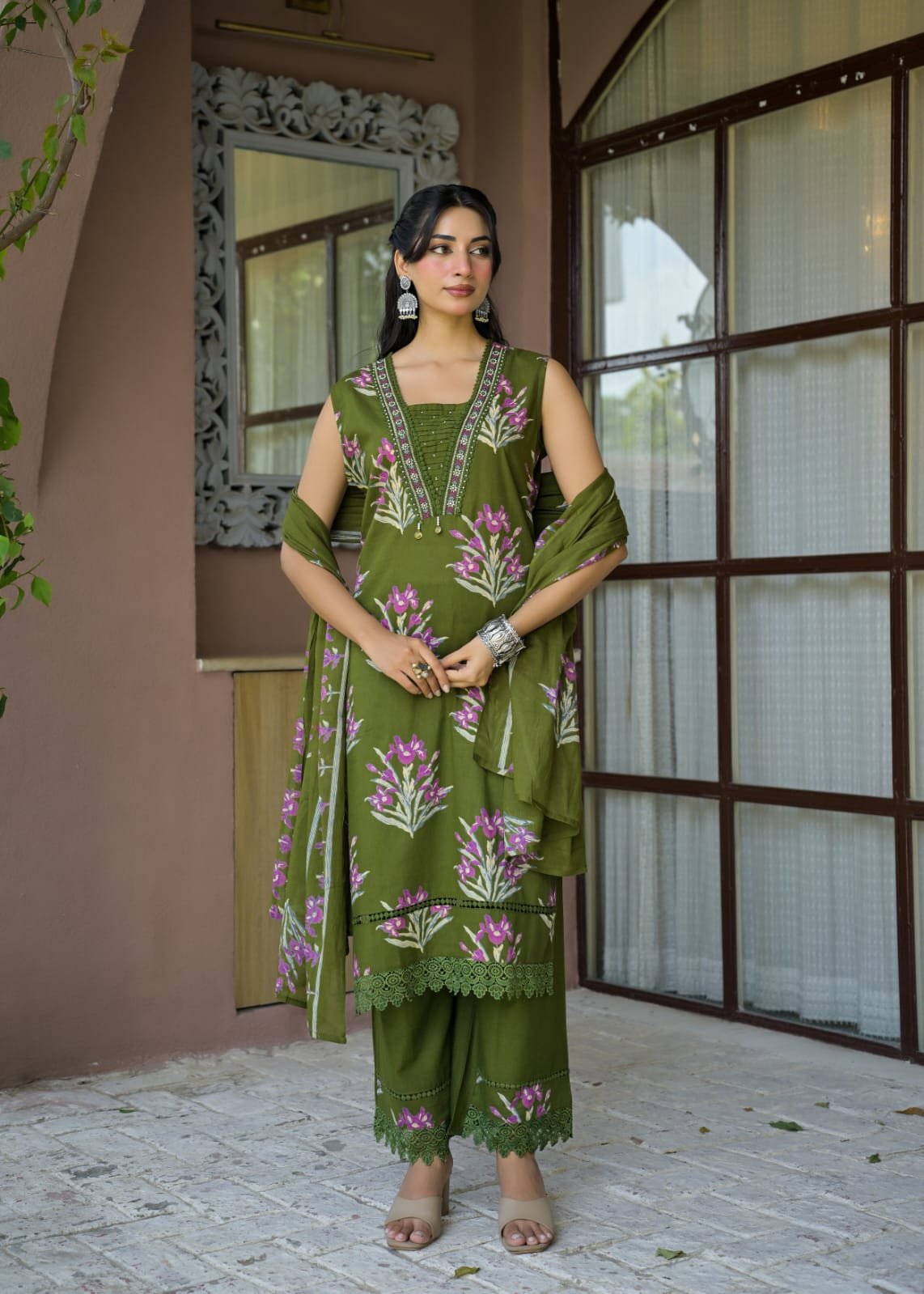 A woman models an olive green three-piece salwar kameez suit with pink and purple floral embroidery, standing in front of a brown stucco wall and window.