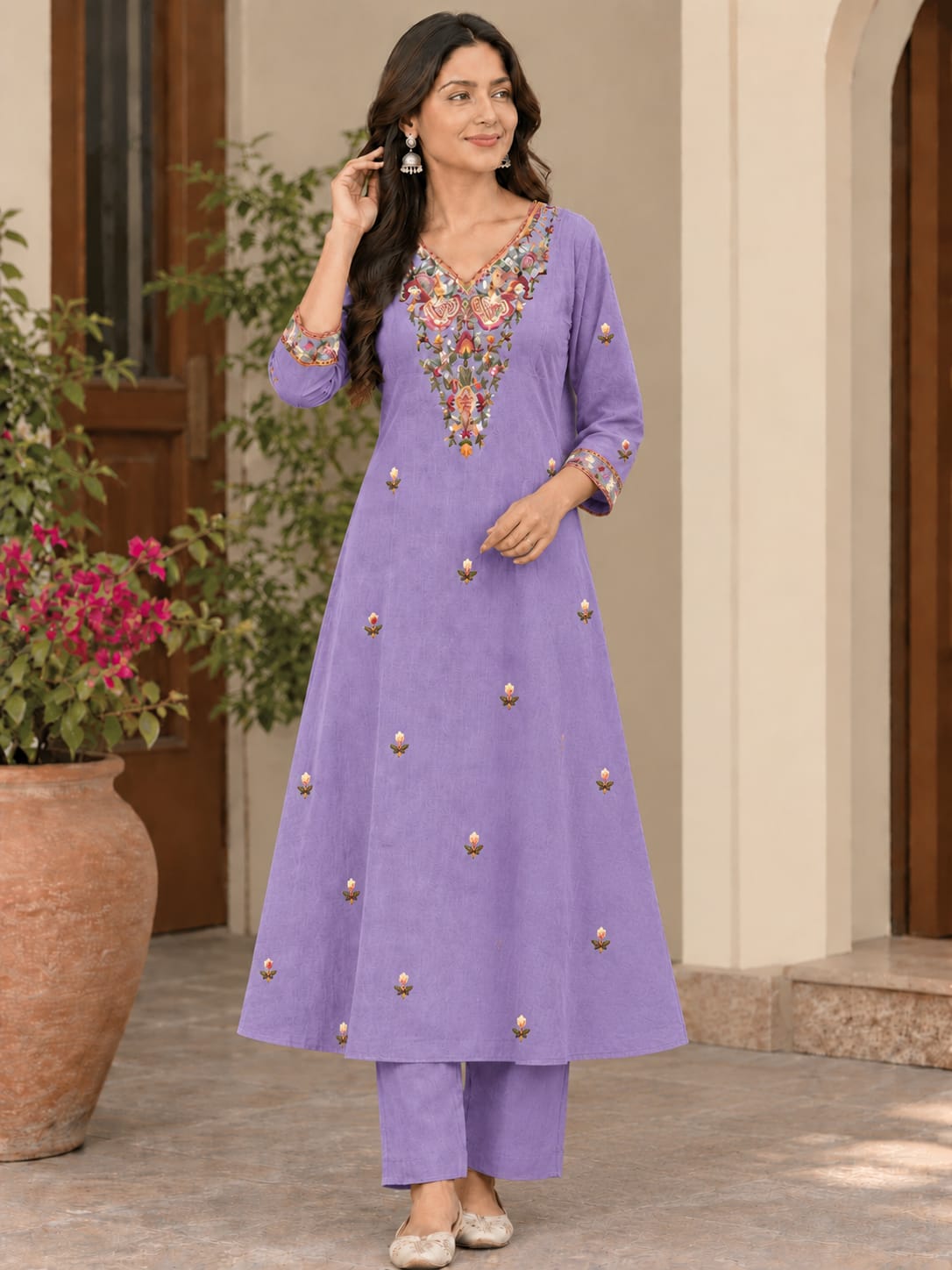 A woman models a lavender purple kurta set with colorful floral embroidery on the neckline and scattered gold-toned embellishments, paired with matching pants, in an outdoor courtyard setting.