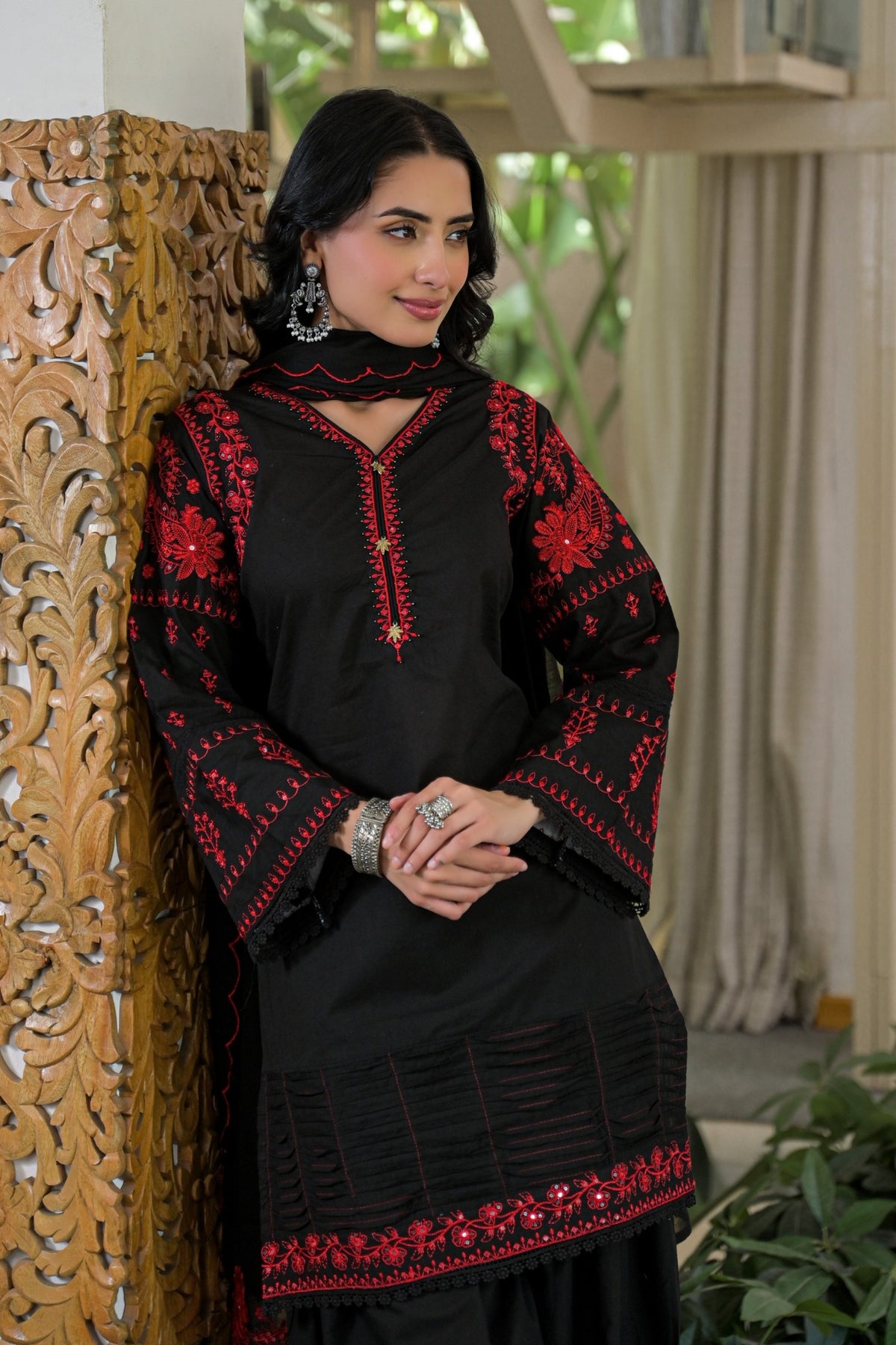 A woman wearing a black embroidered kurta with red floral patterns and a matching black dupatta, leaning against a carved wooden screen in an indoor setting.