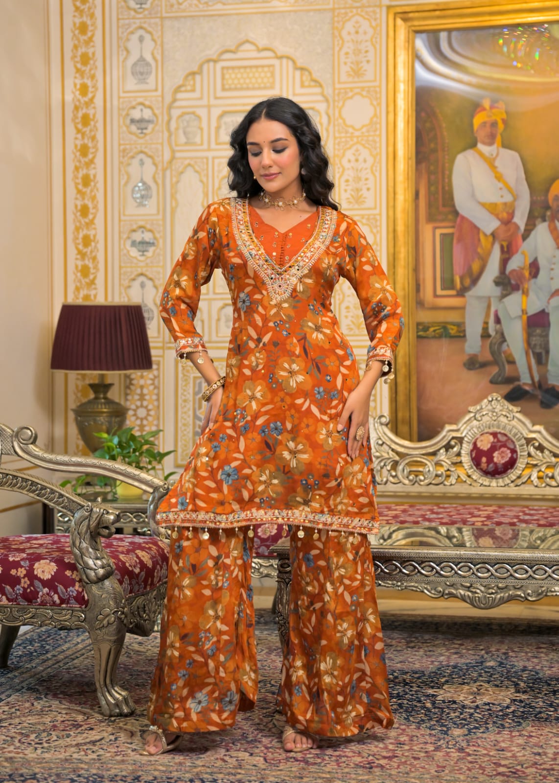 A woman modeling a traditional Indian salwar kameez suit in a highly ornate, vintage-style room. The outfit is a vibrant orange with a detailed floral print in yellow, blue, and green, featuring gold embroidery on the neckline and borders.