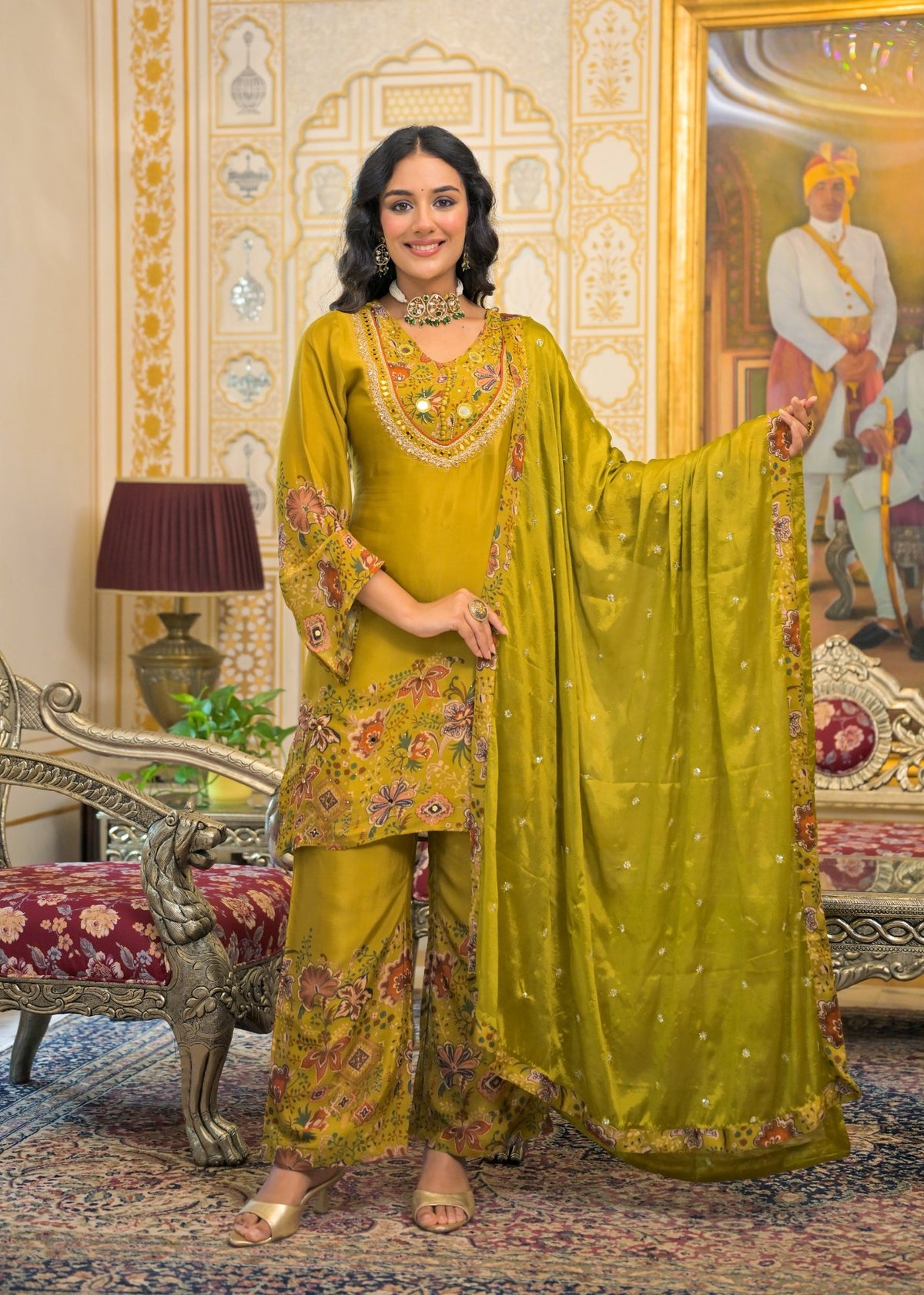 A woman models a traditional Indian salwar kameez suit in olive green with intricate floral embroidery, paired with a matching sheer dupatta, in an ornate indoor setting.