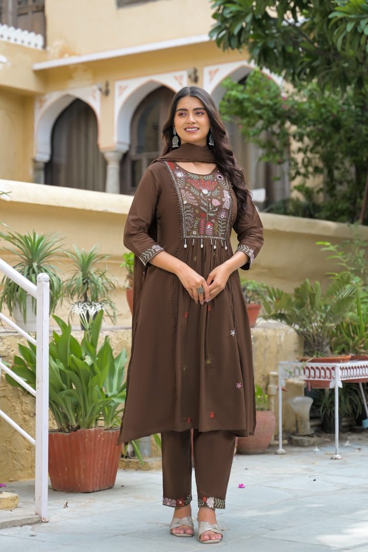 A woman modeling a brown three-piece kurta set with matching pants and dupatta, featuring a heavily embroidered neckline, standing in an outdoor courtyard setting.