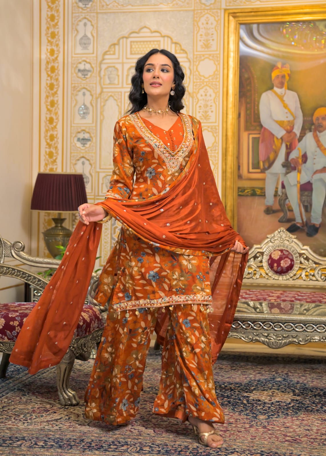 A woman models a traditional Indian salwar kameez suit in a richly decorated palace-like room. The outfit consists of an orange-brown printed kurta and wide-leg pants with a matching printed dupatta.