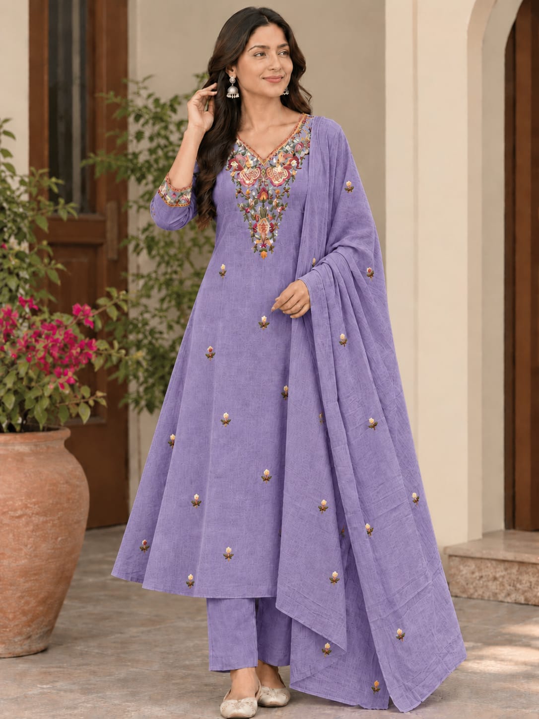 A woman models a lavender purple traditional Indian outfit consisting of a long kurta with colorful floral embroidery on the neckline, matching trousers, and a draped dupatta decorated with small metallic gold buttons, set against an outdoor courtyard background.