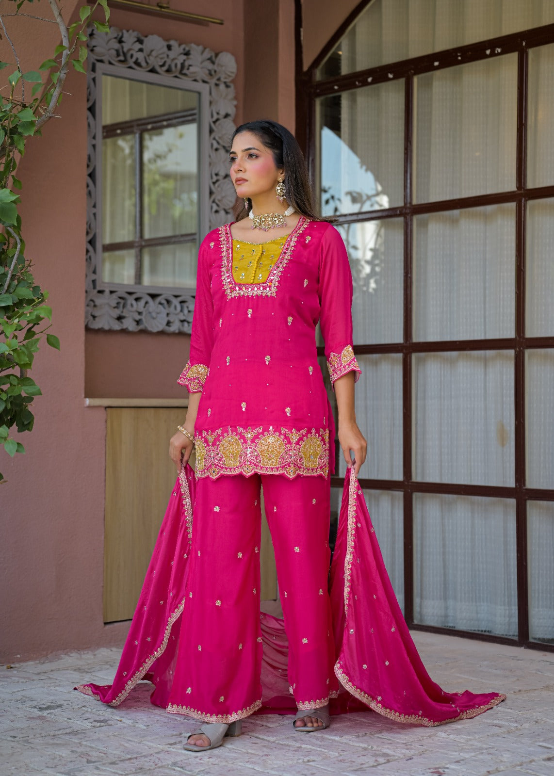 A woman models a bright pink traditional South Asian outfit with gold embroidery, matching wide-leg pants, and a sheer dupatta, standing in front of a pink stucco building with a window.