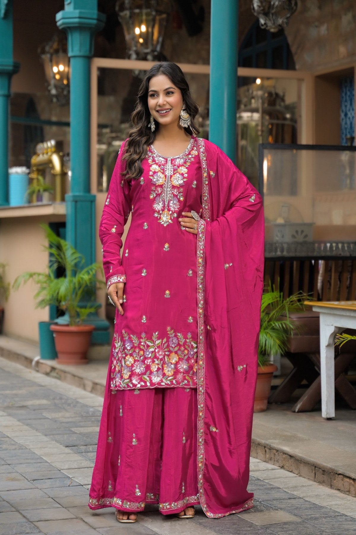 A woman models a bright magenta pink traditional Indian salwar kameez suit with intricate gold and multicolor floral embroidery, paired with a matching dupatta, set against an outdoor architectural background with teal pillars.