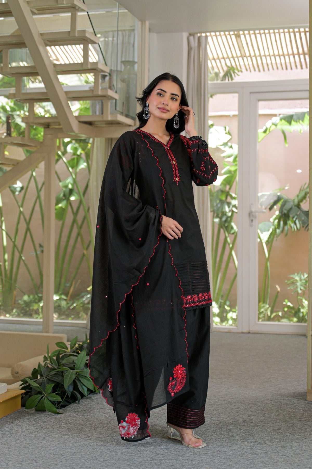 A woman models a black traditional South Asian outfit with red embroidered details, consisting of a long kurta, matching pants, and a draped dupatta, in a modern indoor setting with a staircase and glass doors.