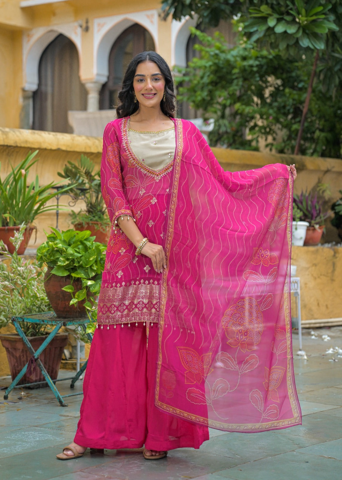 A woman models a vibrant pink traditional Indian salwar kameez suit with a matching printed dupatta in an outdoor courtyard setting.