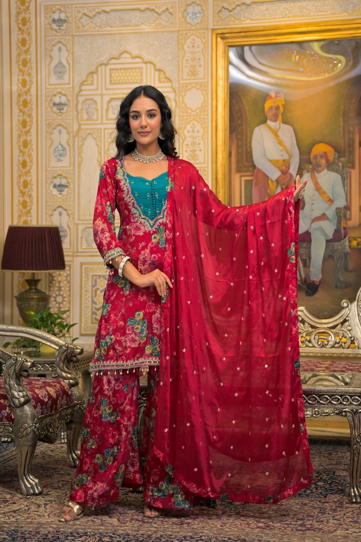 A woman models a traditional Indian salwar kameez suit in a richly decorated palace-like room. The outfit features a deep red floral printed kurta and matching trousers, a teal embroidered kameez bodice, and a matching red dupatta with gold zari embroidery.