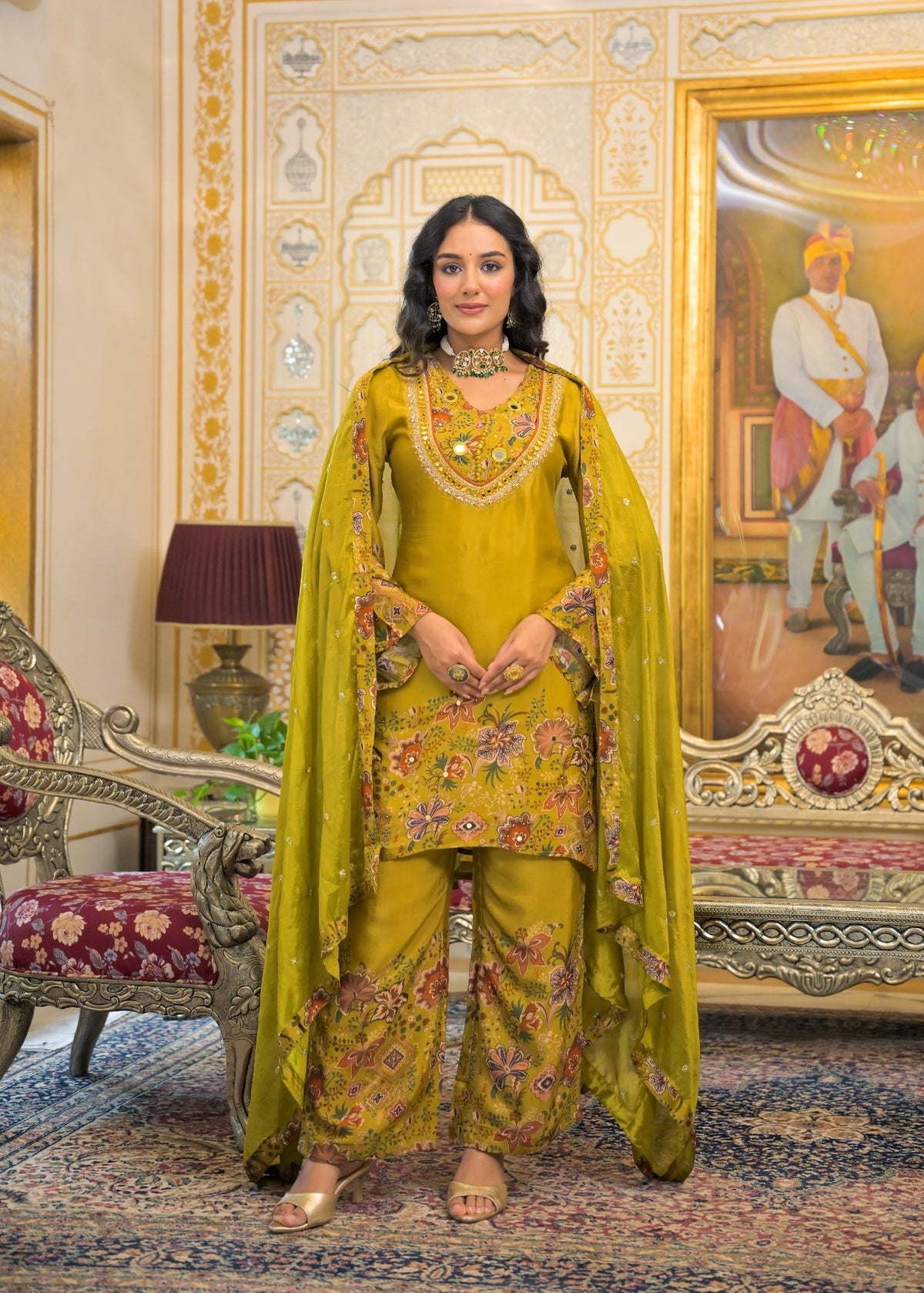 A woman stands in an ornate traditional room wearing a bright yellow-green embroidered salwar kameez suit with a matching dupatta. The outfit features floral embroidery in pink, gold, and green, paired with a heavily embellished gold necklace and matching jewelry.