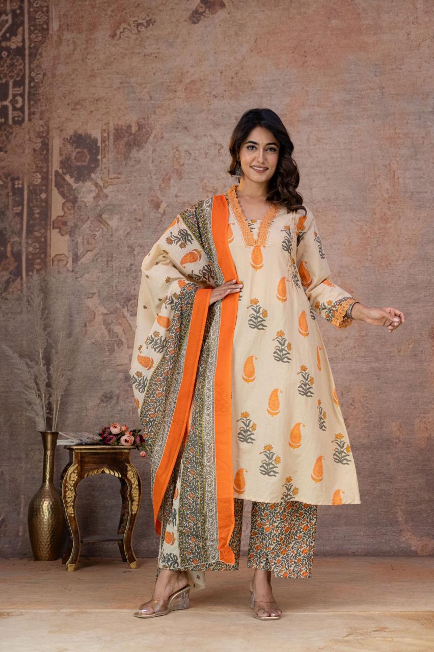 A woman models a traditional Indian outfit consisting of a cream-colored kurta with green leaf prints and green patterned sleeves, paired with matching green patterned pants, set against a rustic textured wall backdrop.