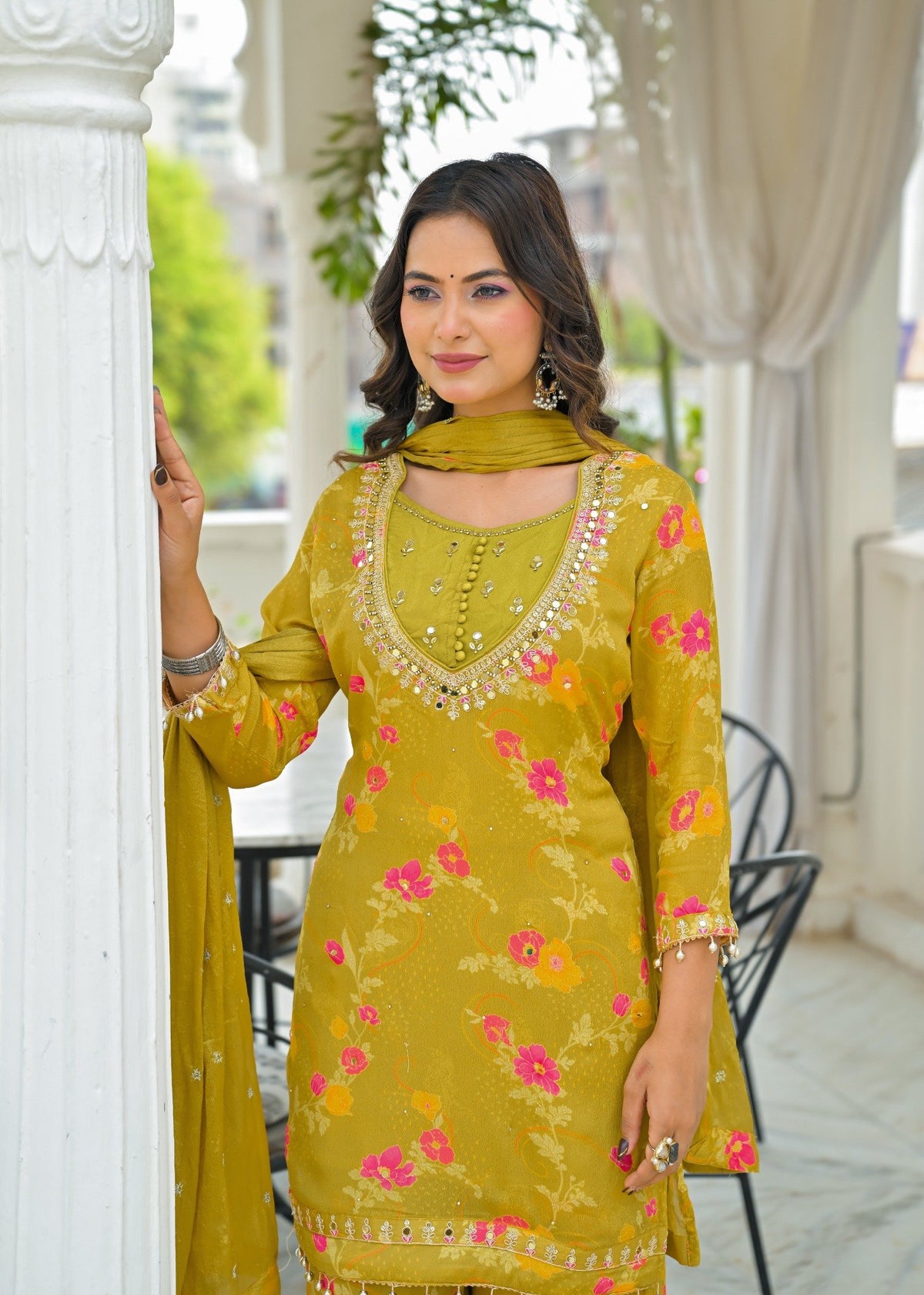A woman models a yellow-green floral salwar kameez suit with a matching dupatta, standing in a white-walled corridor with columns.