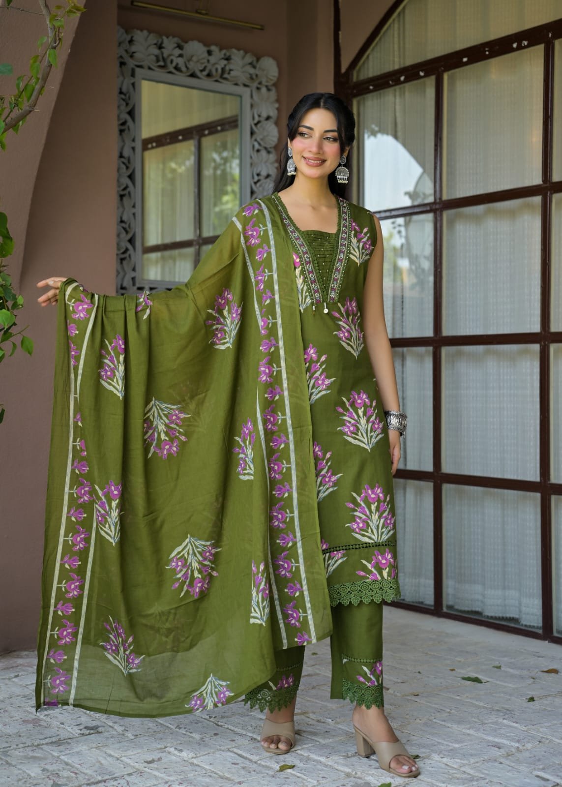 A woman models an olive green three-piece traditional outfit with pink and white floral embroidery, including a kurta, pants, and dupatta, set against a warm-toned architectural background.