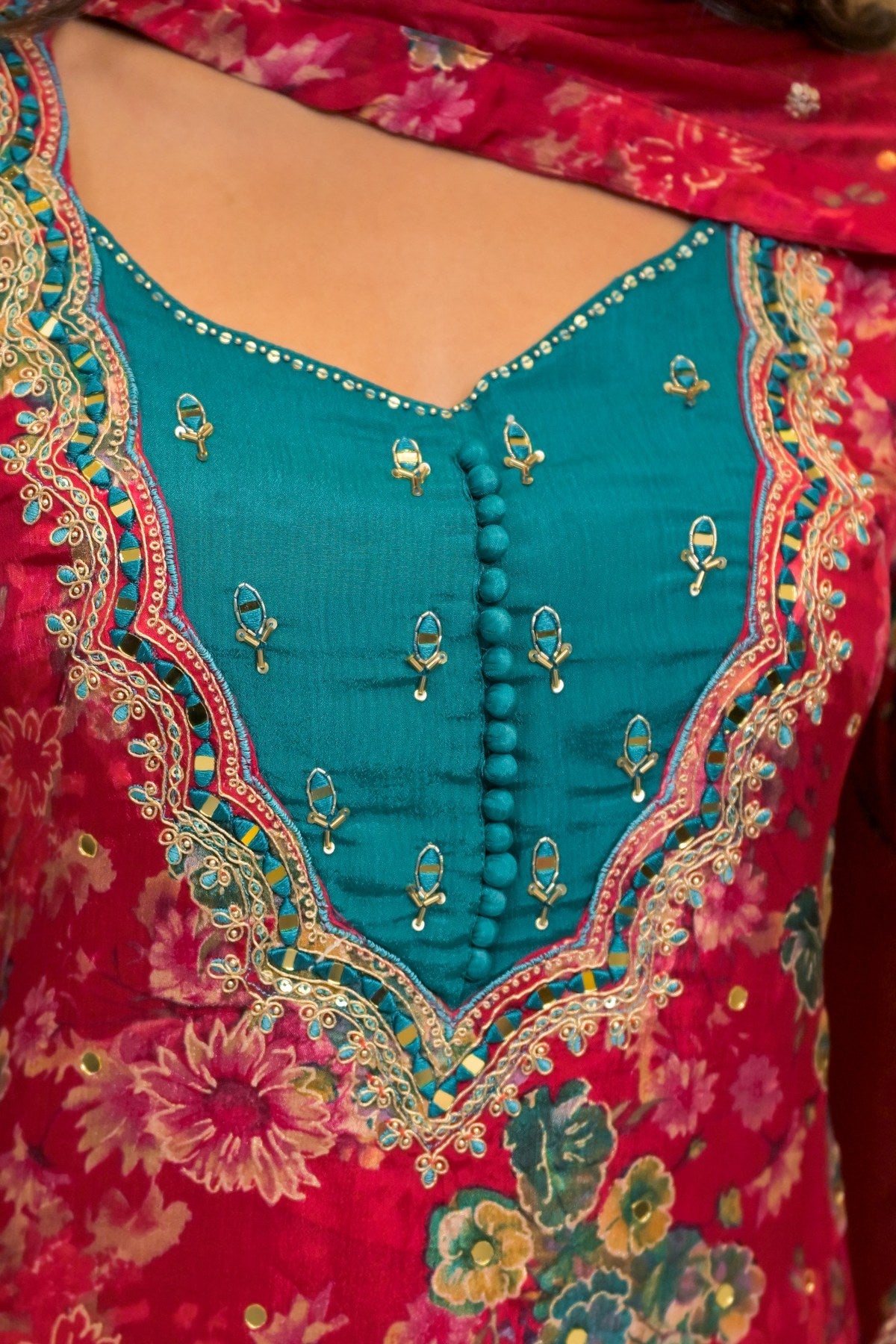 Close-up of a traditional Indian salwar kameez garment featuring a vibrant teal embroidered yoke with gold mirror work and a bright pink floral printed fabric body.