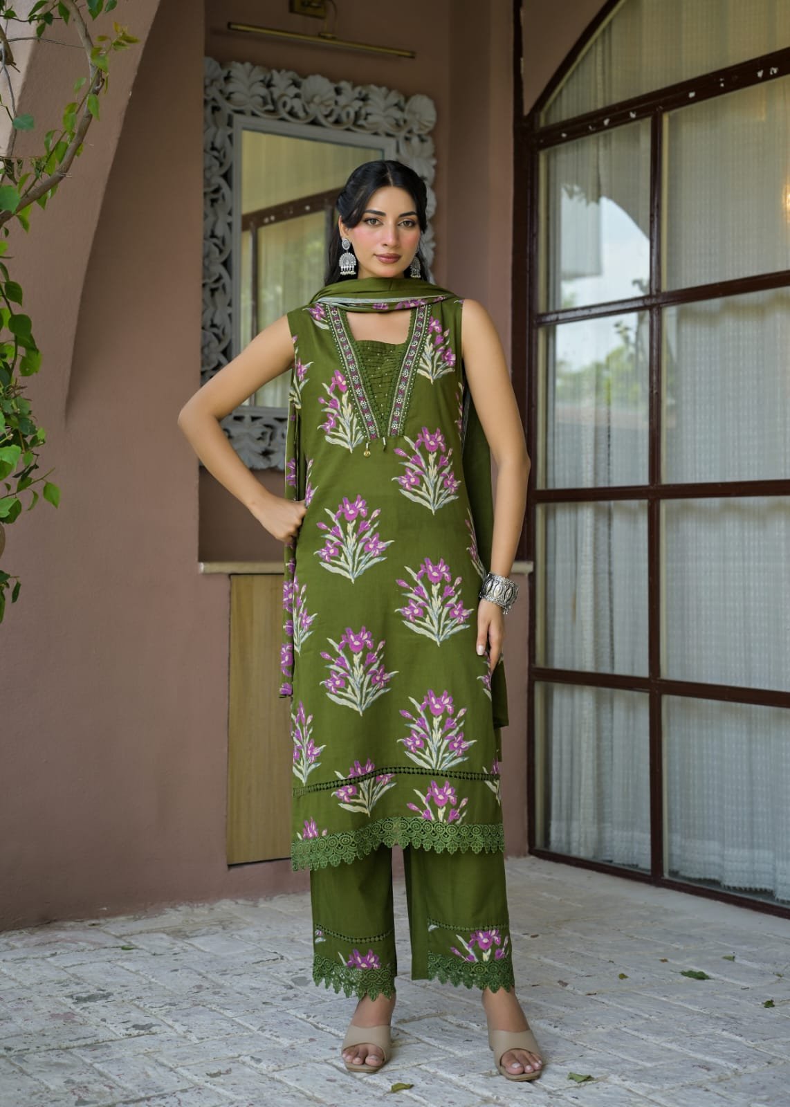 A woman models an olive green three-piece traditional outfit with pink floral embroidery, standing on a stone patio in front of a brown stucco wall and glass window.