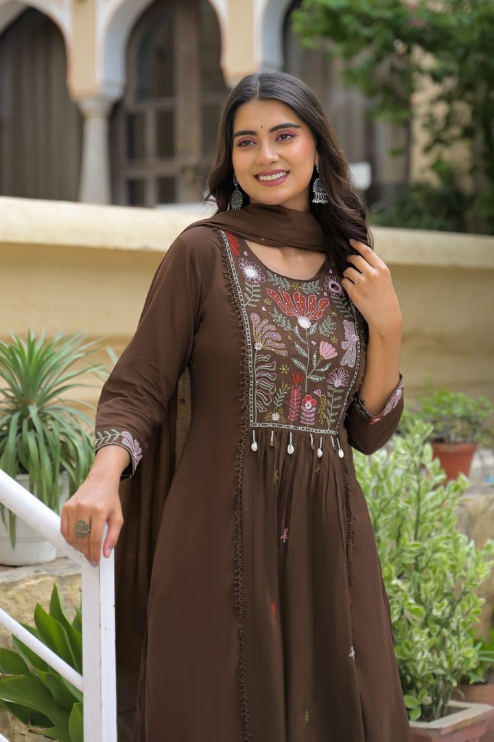 A woman wearing a brown long kurta with floral embroidery on the chest and sleeves, paired with a matching brown dupatta, standing outdoors in front of a building with arches and potted plants.