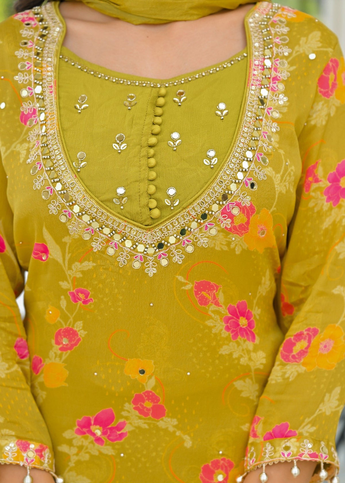 A close-up of a mustard yellow traditional outfit with pink and orange floral patterns, decorated with gold and silver mirror work and bead borders on the neckline and cuffs.