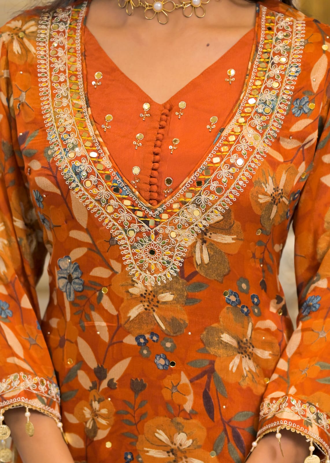 Close-up of a person wearing an orange floral embroidered tunic with a V-neck, heavily decorated with gold-toned mirror work and metallic embellishments.