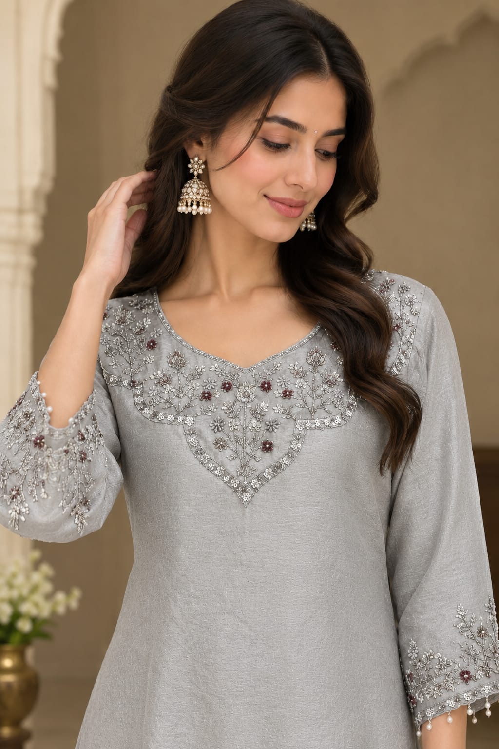 A woman wearing a light gray three-quarter sleeve kurta with intricate silver and maroon beadwork around the V-neck and sleeve cuffs, styled with jhumka earrings.