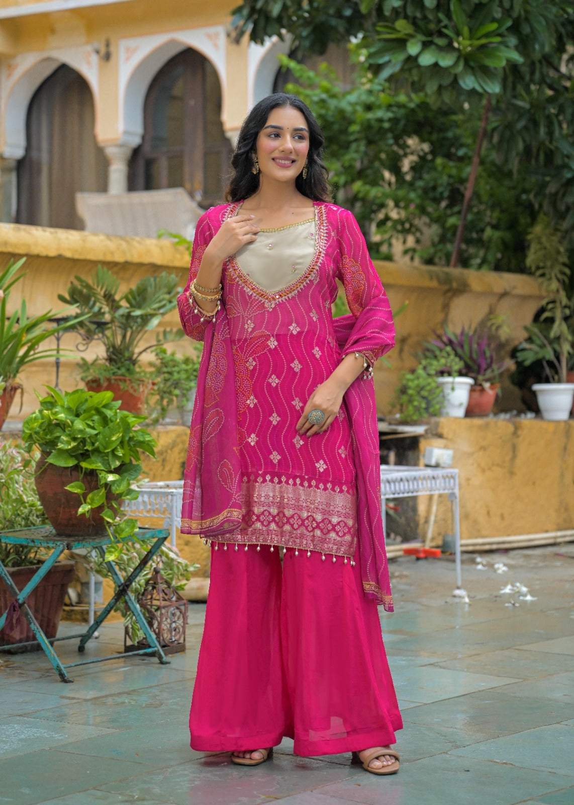 A woman models a bright pink Indian ethnic salwar kameez suit with a beige embroidered inner panel, wide-leg trousers, and a matching dupatta in an outdoor courtyard setting.