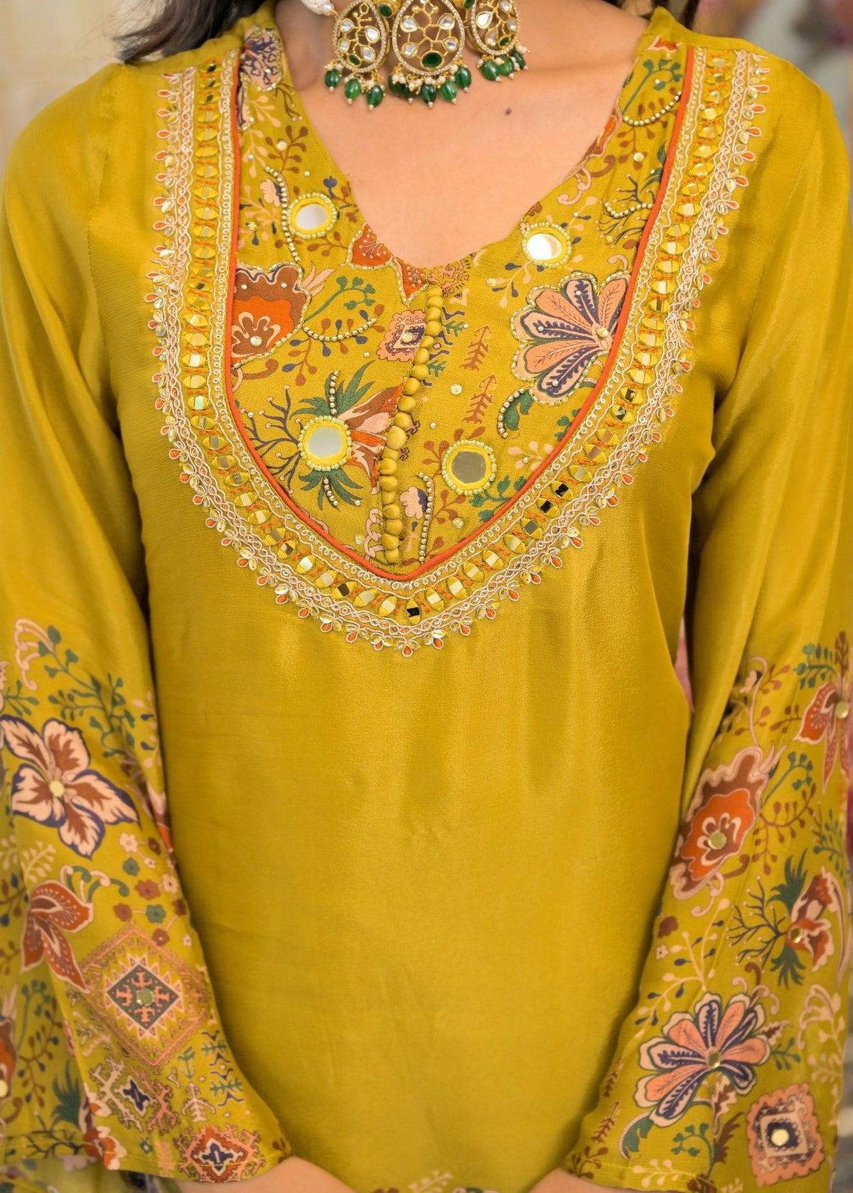 A close-up of a mustard yellow embroidered kurta featuring a V-neckline with intricate floral and geometric patterns, mirror work, and gold zari detailing, along with matching embroidered sleeves.