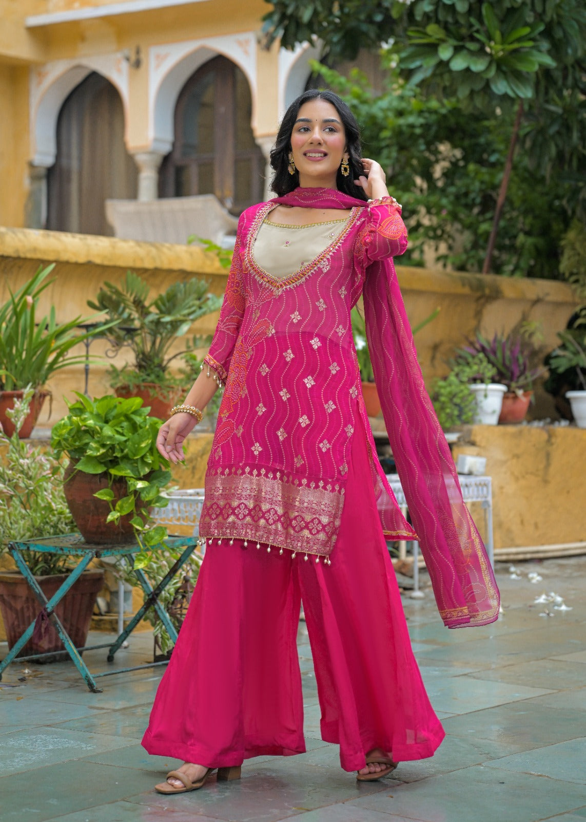 A woman models a vibrant magenta pink Indian ethnic salwar kameez suit with a beige inner kameez and gold embroidery, set against a background of a courtyard with potted plants and a yellow building.