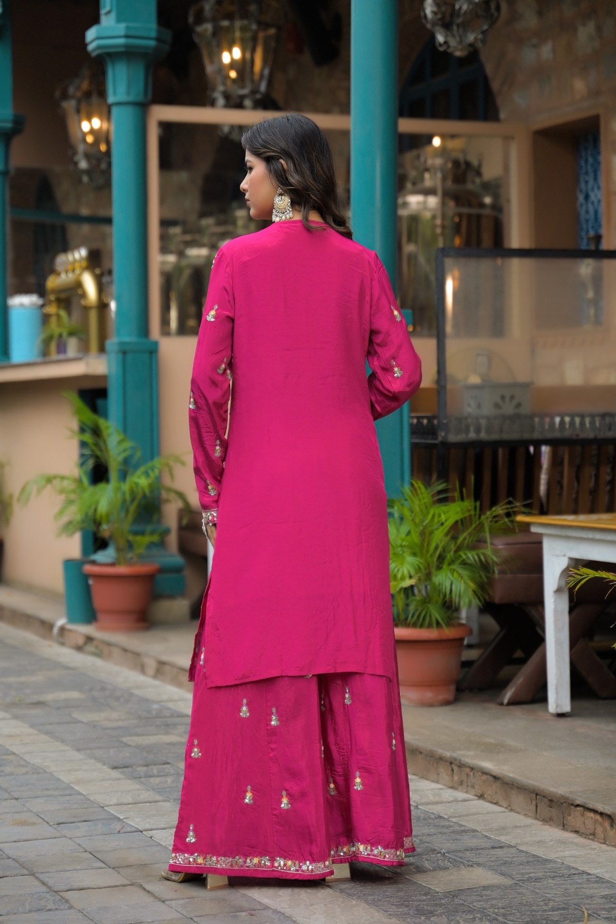 A woman wearing a bright pink traditional outfit with gold embroidery, photographed from behind in an outdoor courtyard setting.