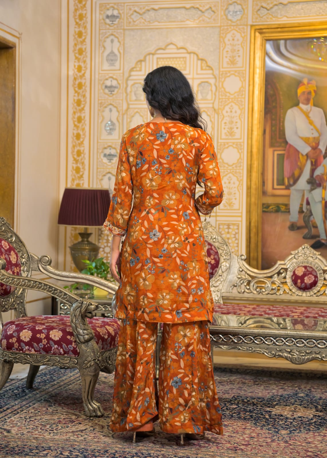 A woman stands with her back to the camera wearing a matching orange floral printed kurta and trouser set in an ornate, traditional Indian palace interior with gold-framed paintings and carved furniture.