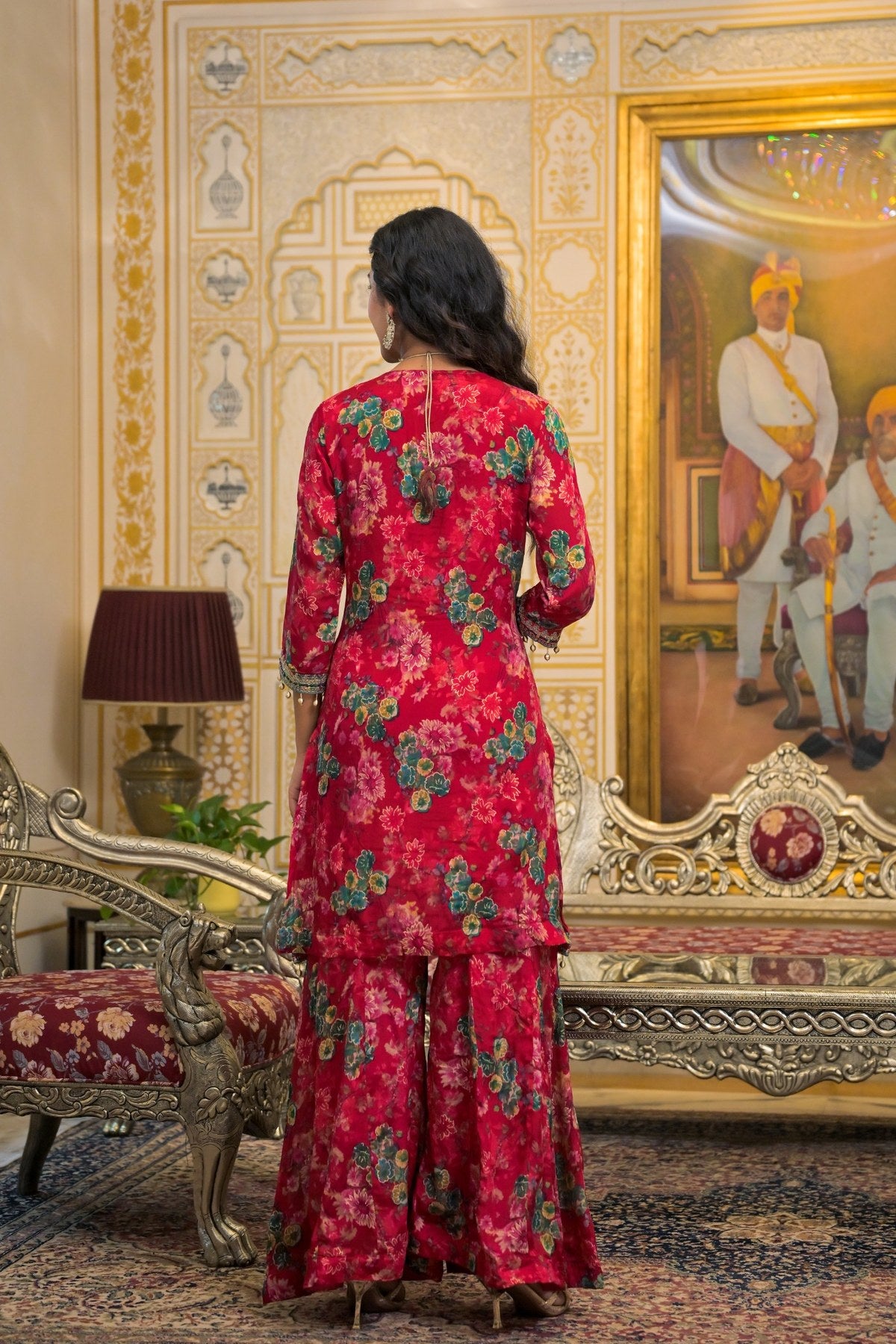 A woman stands with her back to the camera wearing a red floral printed traditional outfit with matching pants in an ornate, vintage-style room with gold furniture and a large framed painting.